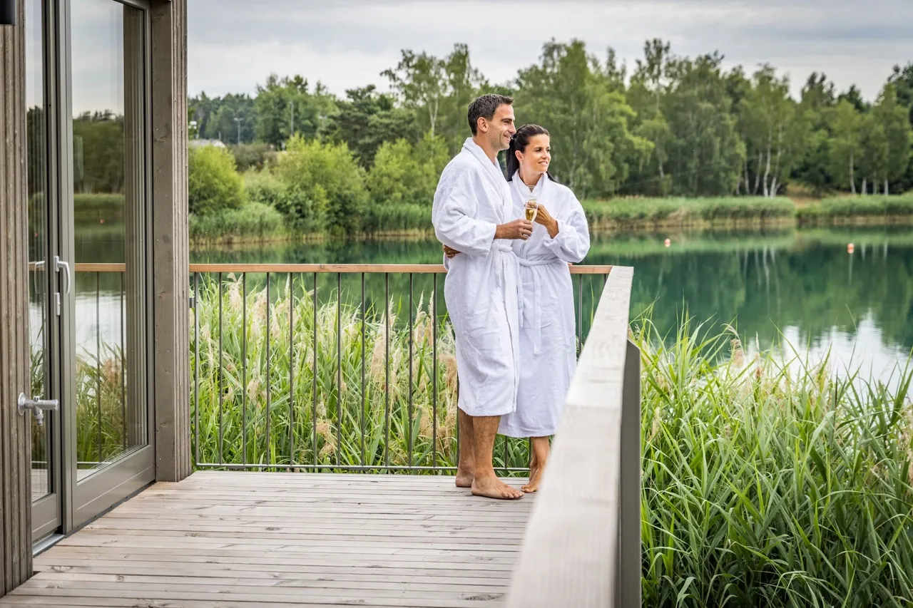Couple in white bathrobes with sparkling wine on a wooden terrace overlooking a lake