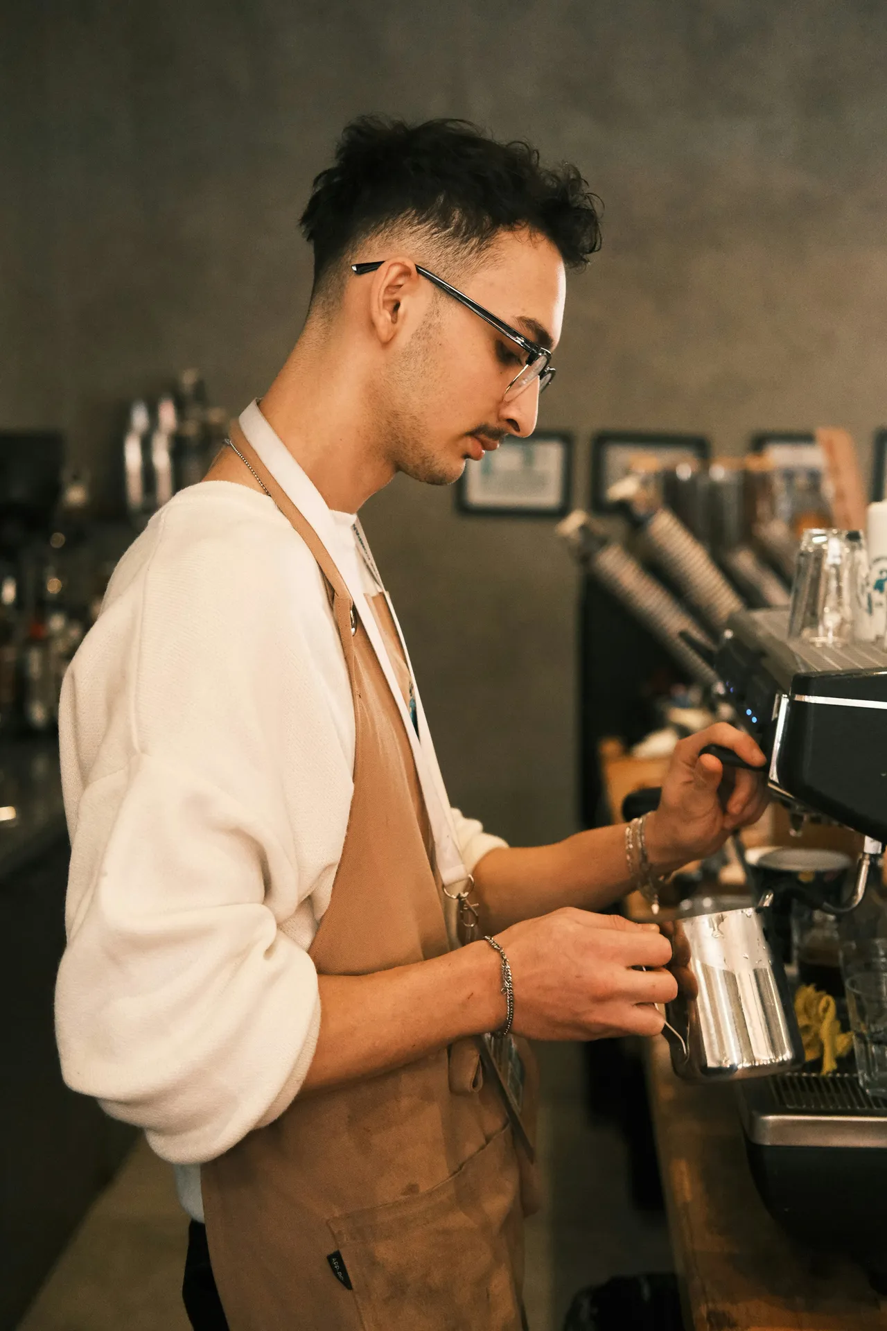 Male barista in cream-colored shirt and tan apron operating an espresso machine, with blurred coffee equipment and bottles visible in the background.