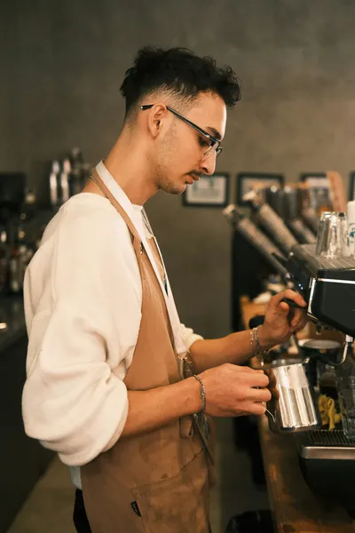 Male barista in cream-colored shirt and tan apron operating an espresso machine, with blurred coffee equipment and bottles visible in the background.