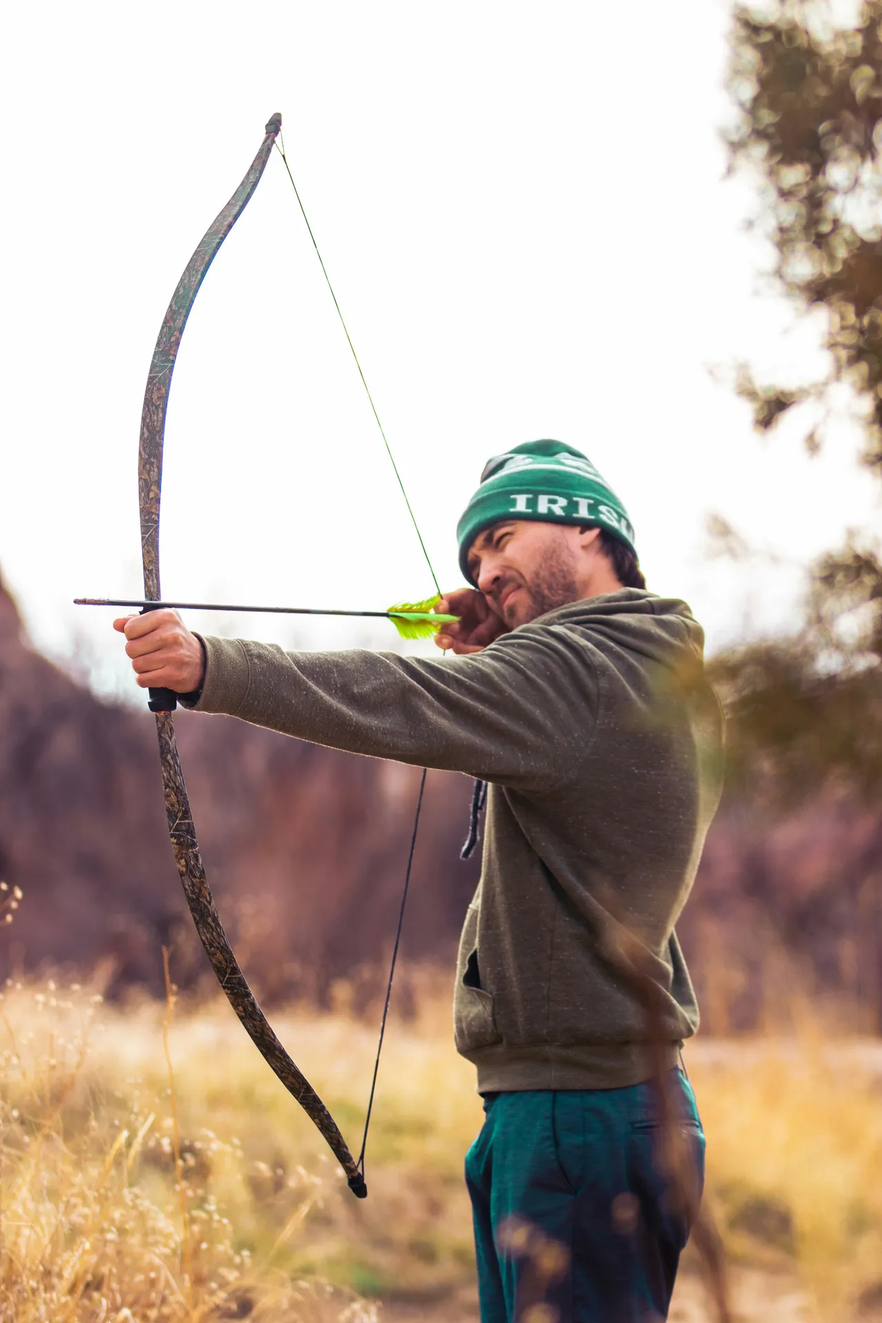 A man wearing a green beanie and hoodie drawing a camo-patterned recurve bow with a green-fletched arrow in a grassy field.