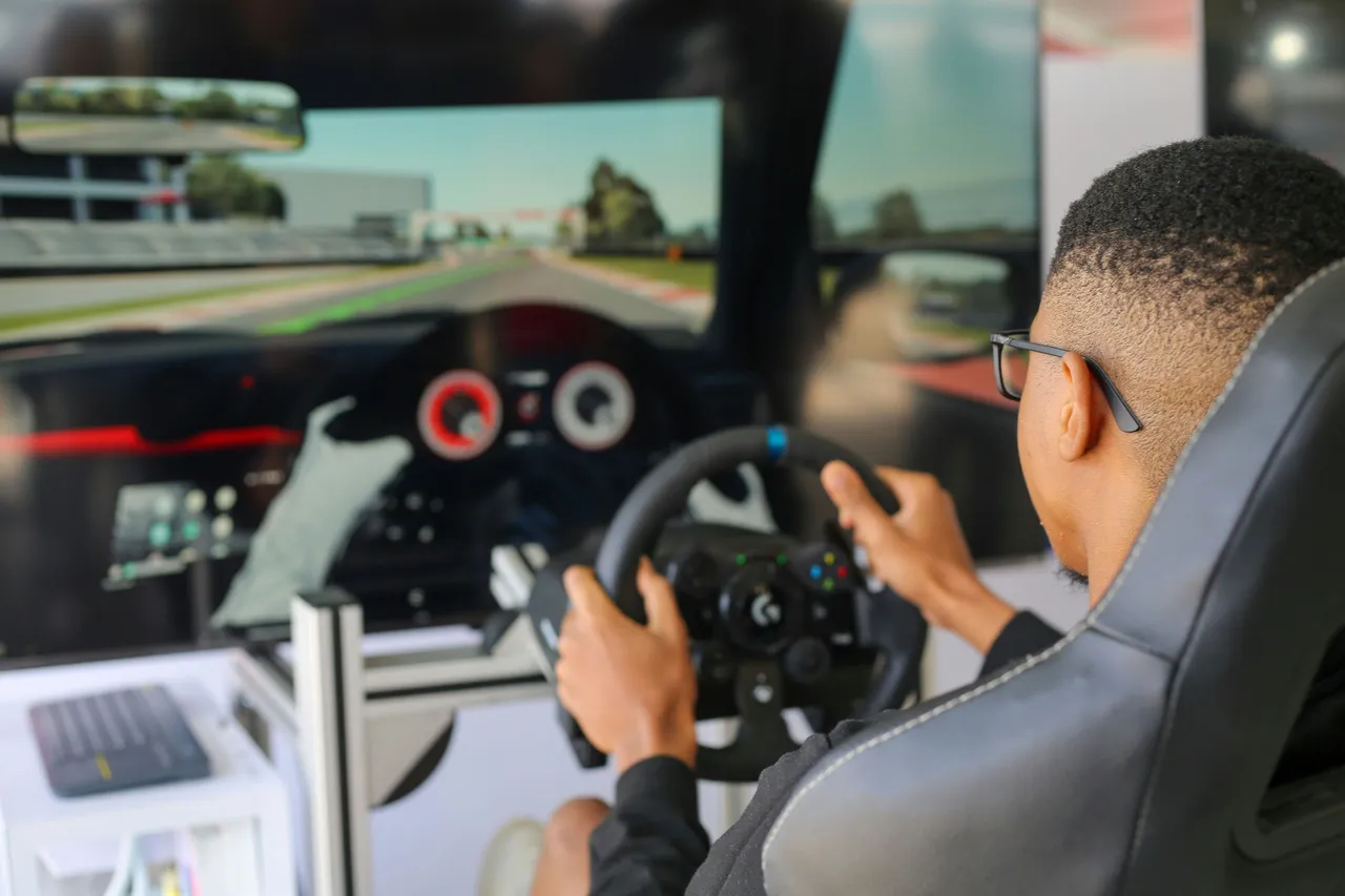 : A man wearing a VR headset sits in a specialized racing simulator cockpit with a steering wheel and pedals, playing a driving game.