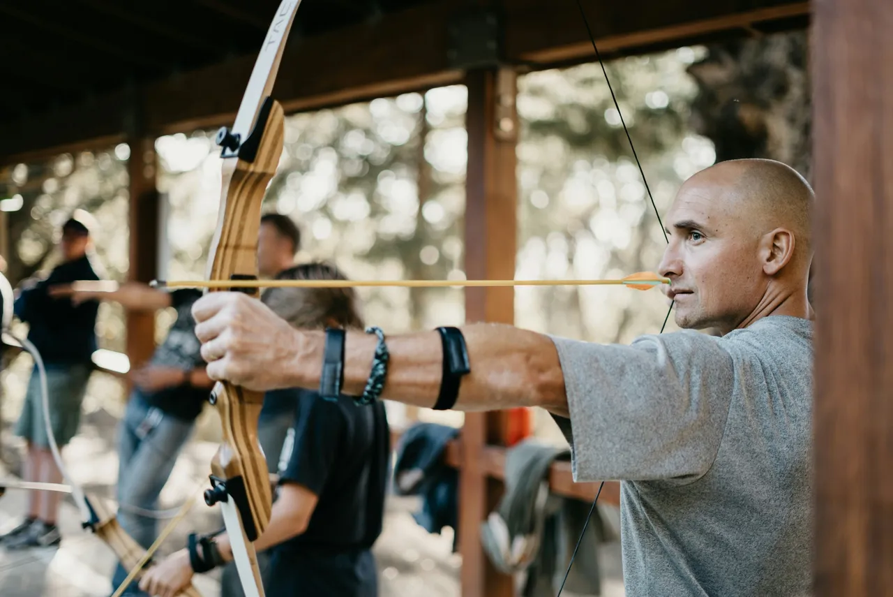 Close-up profile of a bald man drawing a wooden recurve bow with a yellow arrow at a covered archery range.