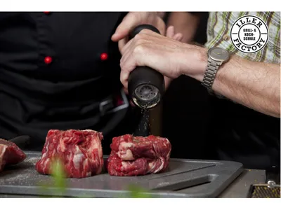Chef seasoning raw beef cuts with a pepper mill at a steel grill station