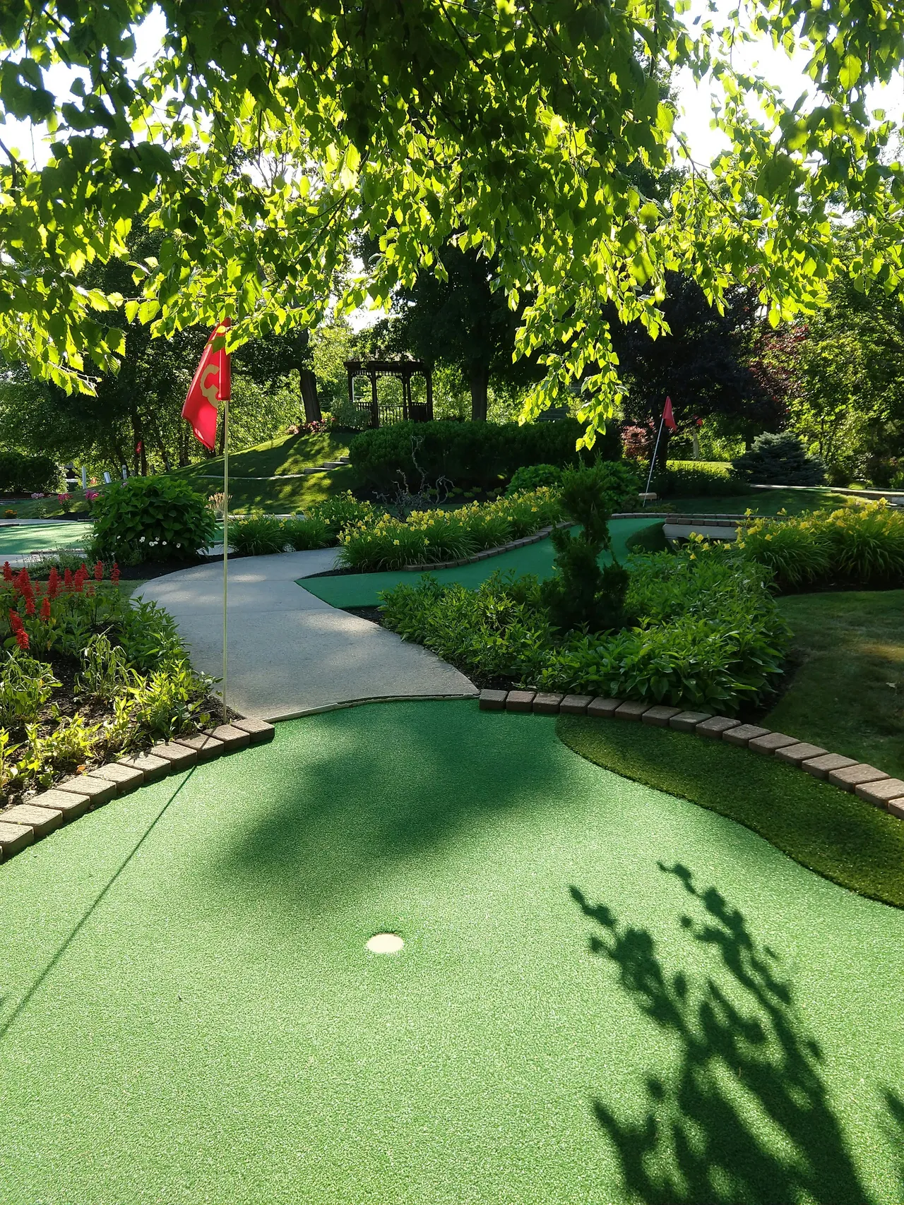 Landscaped minigolf course with a red flag and putting green surrounded by flowering gardens and trees