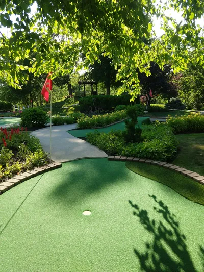Landscaped minigolf course with a red flag and putting green surrounded by flowering gardens and trees