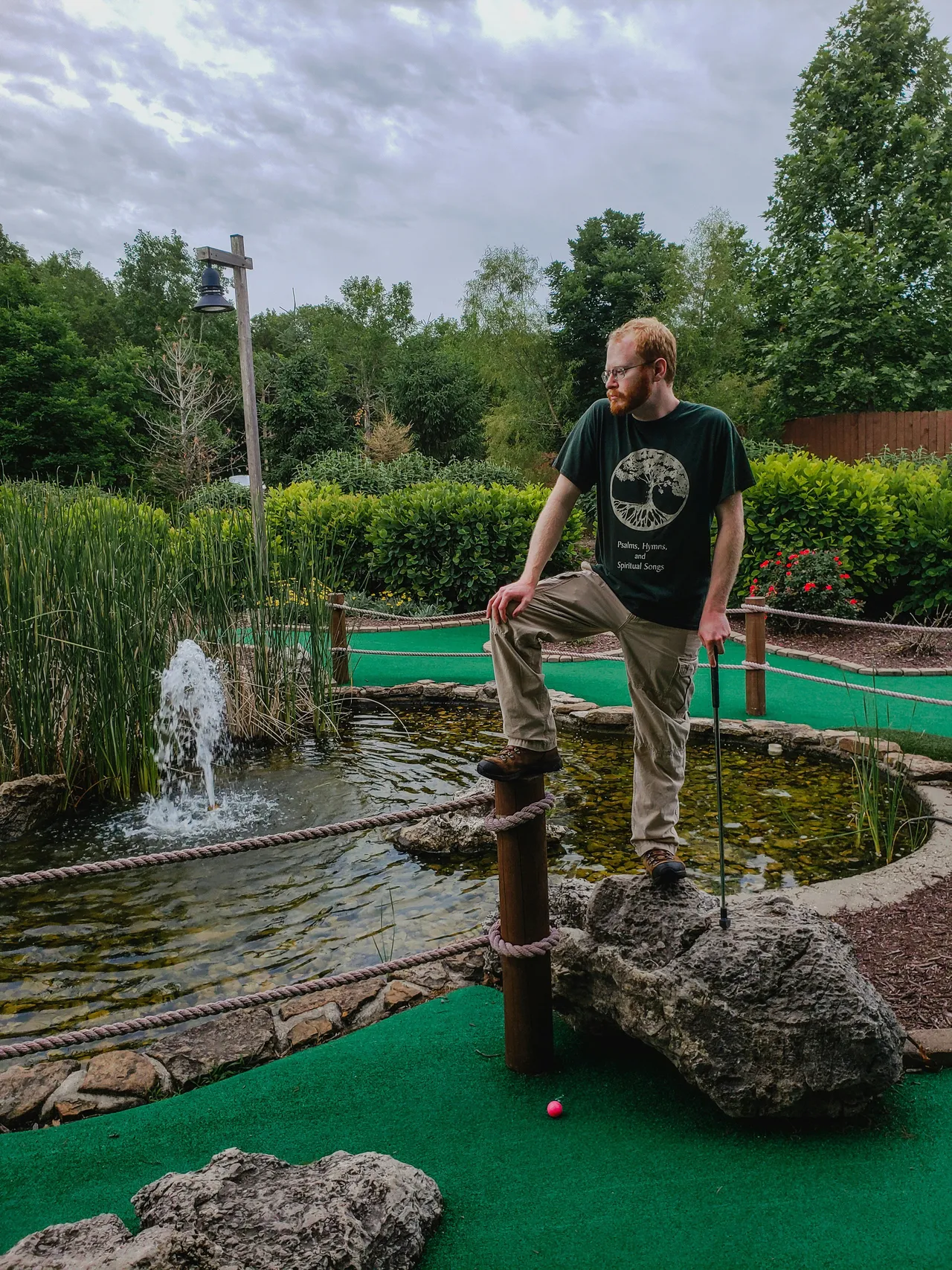 Man balancing on a rock while lining up a minigolf shot beside a pond with a fountain