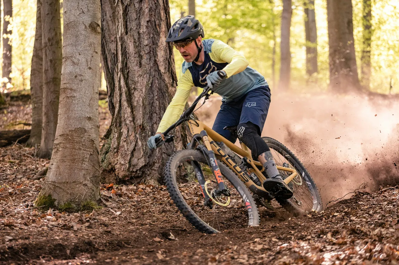 A close-up action shot of a mountain biker kicking up dirt while navigating a sharp, dusty turn in a forest.