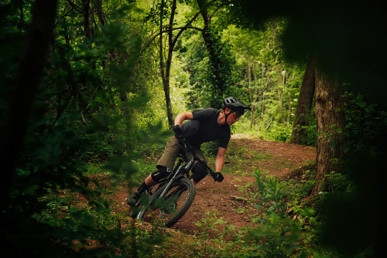 A mountain biker in a dark t-shirt and knee pads leans into a fast, dusty turn on a wooded dirt path.