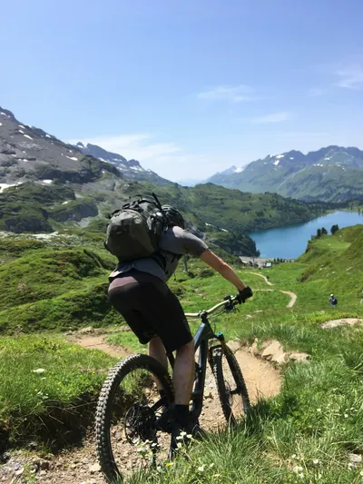 Rear view of a mountain biker riding down a singletrack trail overlooking a bright blue alpine lake and green mountain peaks.
