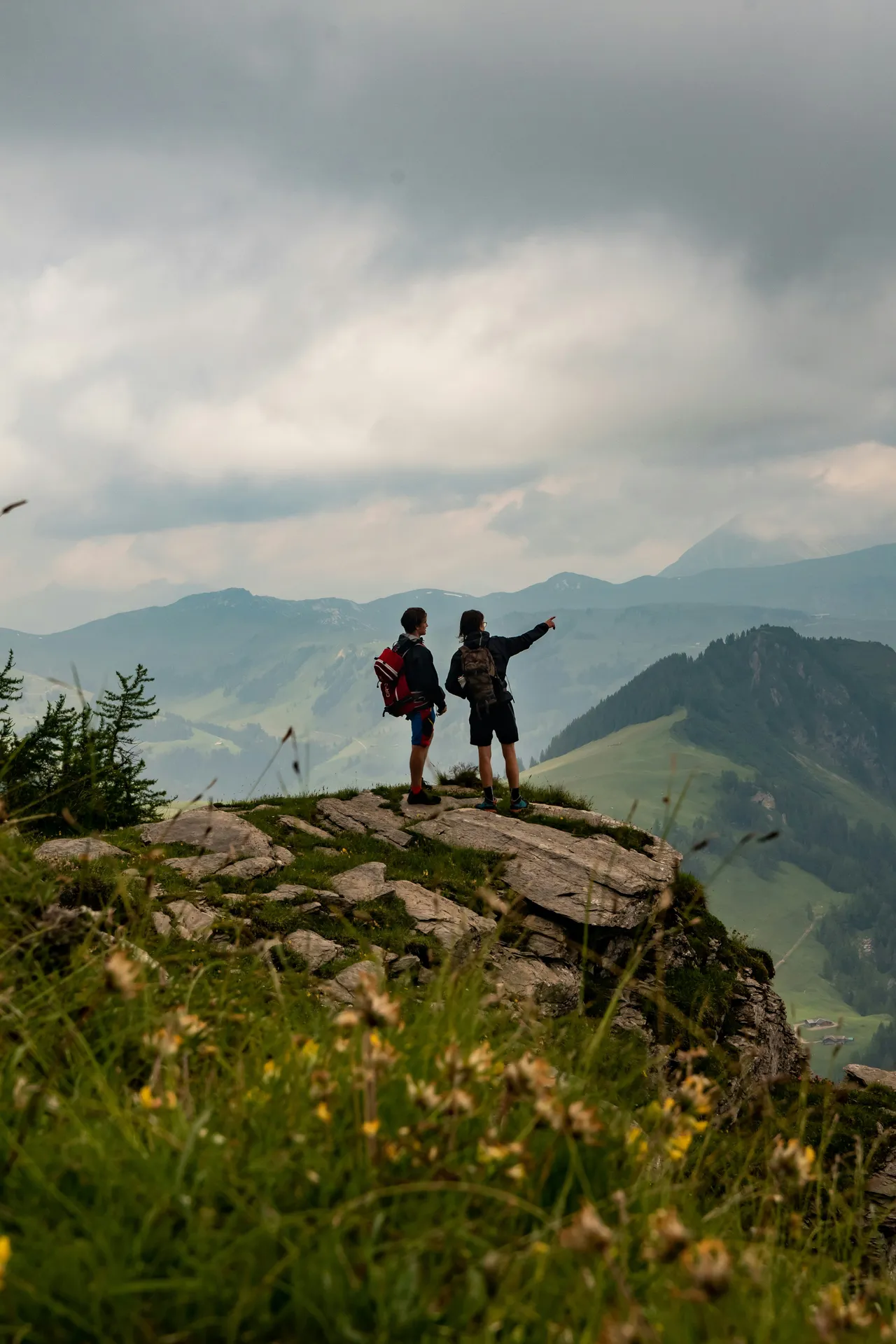 Two hikers with backpacks standing on a rocky summit, pointing toward distant mountain peaks