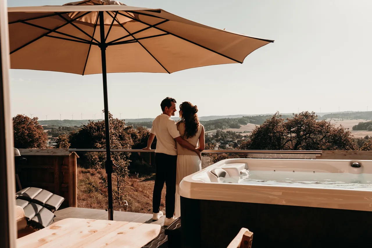 Couple standing on a chalet terrace with hot tub and parasol, looking out over the countryside