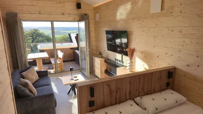 Bright wooden chalet living area with sofa, TV and double bed, opening onto a balcony with hot tub and countryside view