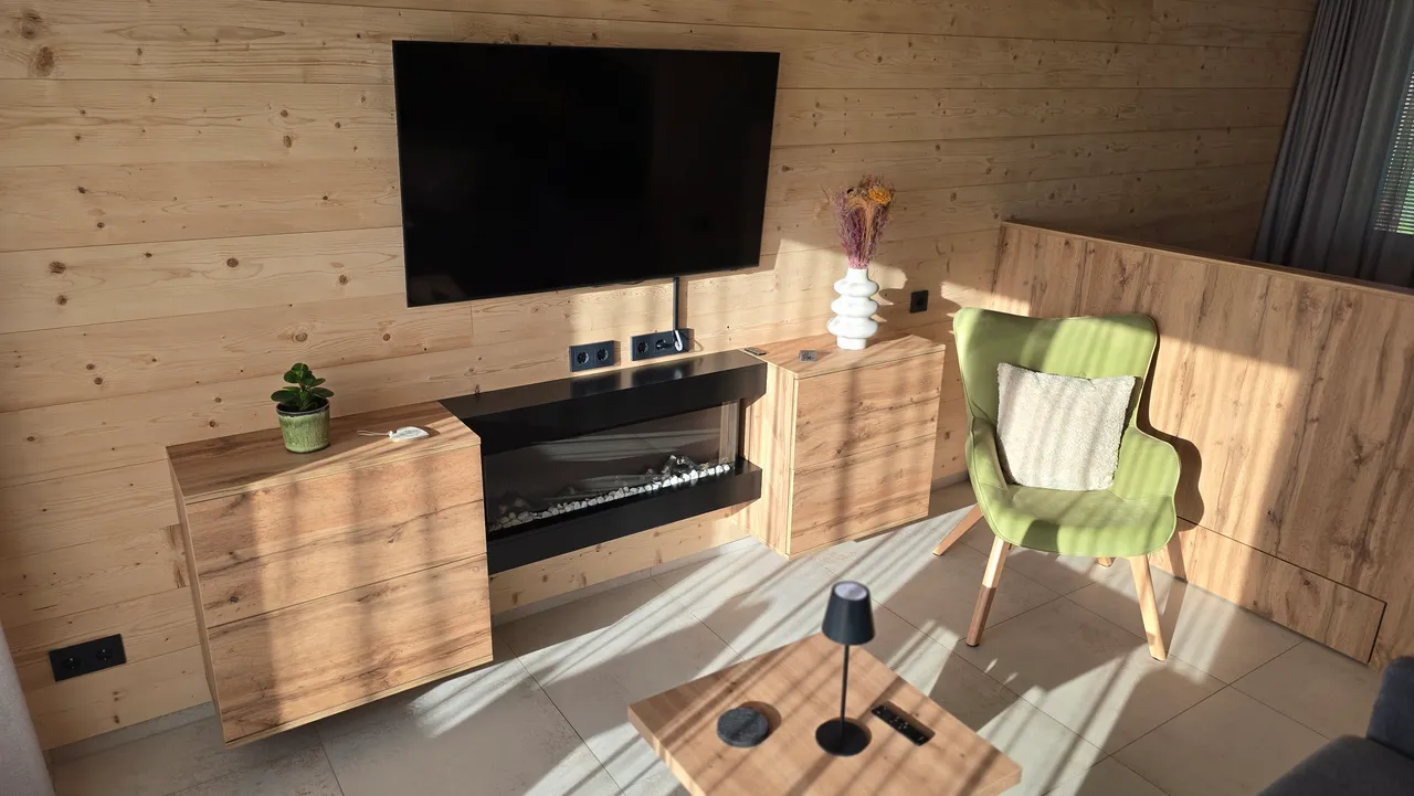 Sunlit chalet living room with wall-mounted TV, electric fireplace and green armchair