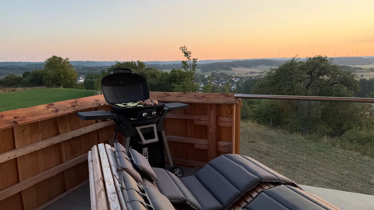 Chalet terrace with grill and cushioned loungers overlooking a green valley at dusk