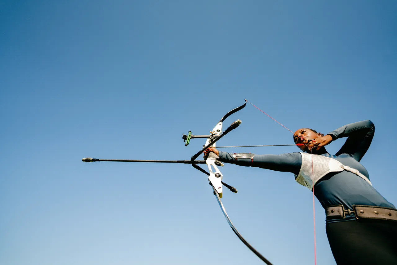 Low-angle shot of a female archer drawing a modern white Olympic recurve bow equipped with stabilizers against a clear blue sky.