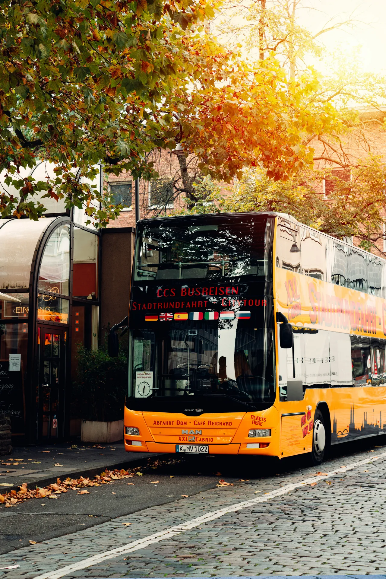 Orange double-decker city tour bus parked on a cobblestone street under autumn trees in Cologne