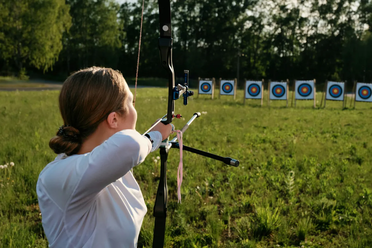 A female archer with a bun aims a compound or modern recurve bow across a wide grassy field toward a row of distant targets.
