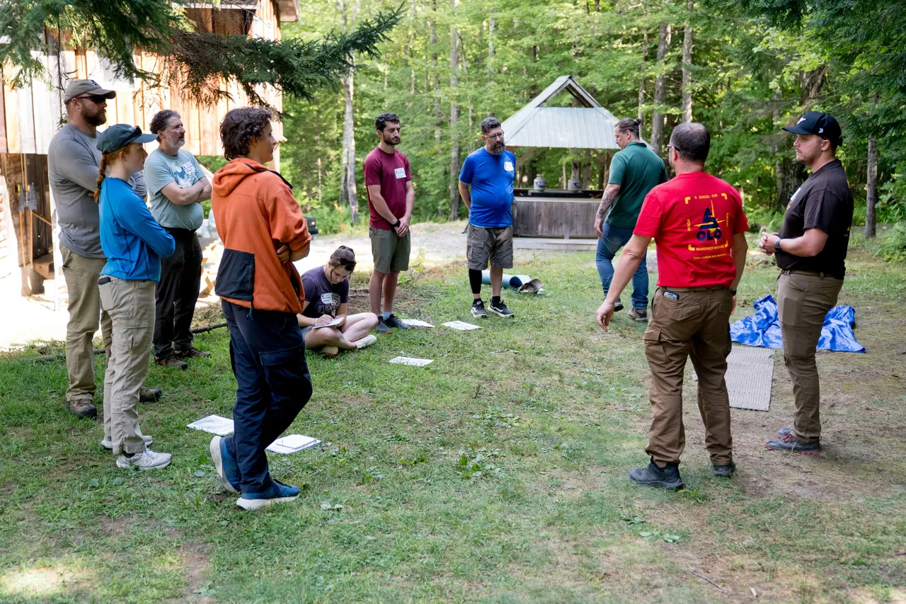 A group of people standing in a circle on a grassy clearing in the woods, listening to instructors during an outdoor survival course.