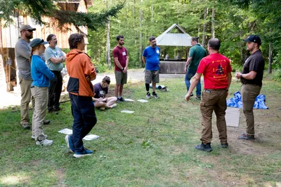 A group of people standing in a circle on a grassy clearing in the woods, listening to instructors during an outdoor survival course.