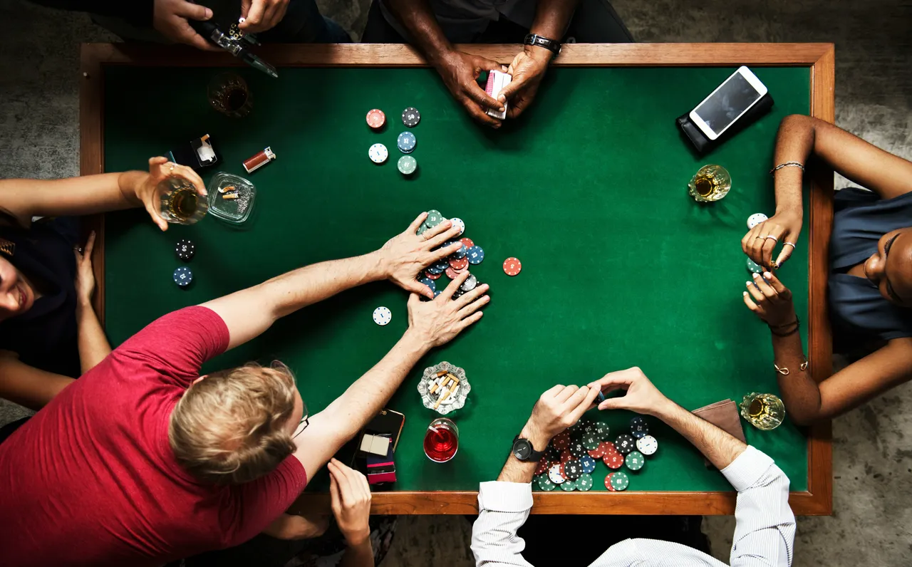 Overhead view of a poker game with players' hands, cards, chips, drinks, and cigarettes on a green table