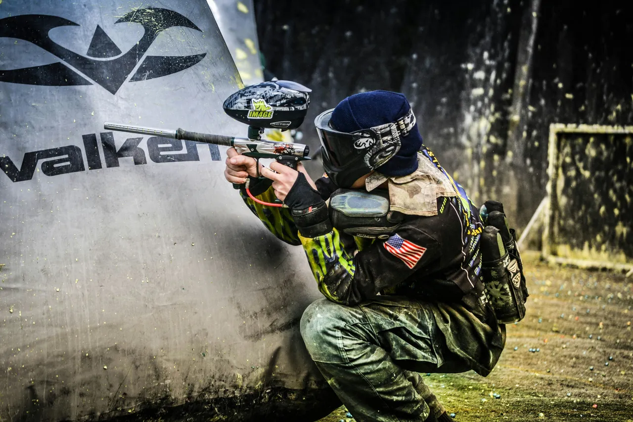 A paintball player in full protective gear and a mask crouching behind an inflatable bunker while aiming a paintball marker.