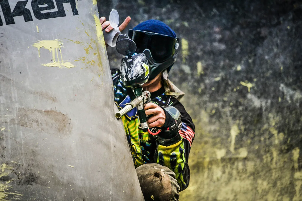 A paintball player in protective gear, a mask, and camouflage clothing running across a dirt field while carrying a paintball marker.