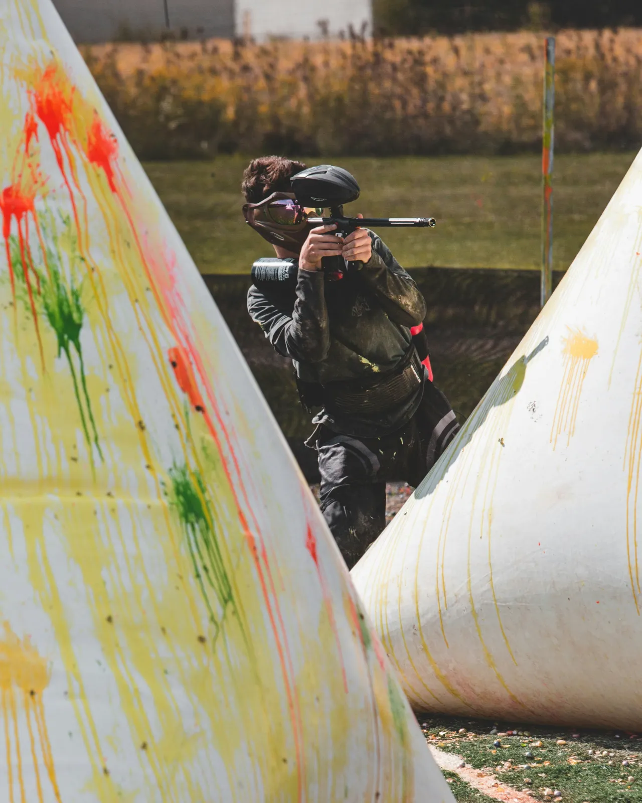 Paintball players dressed in full protective gear, masks, and camouflage hold their markers while navigating an outdoor tactical obstacle field.