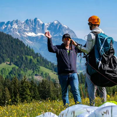 A paragliding instructor checking a student's harness and lines in a wildflower meadow, with the jagged snow-capped Wilder Kaiser peaks in the background