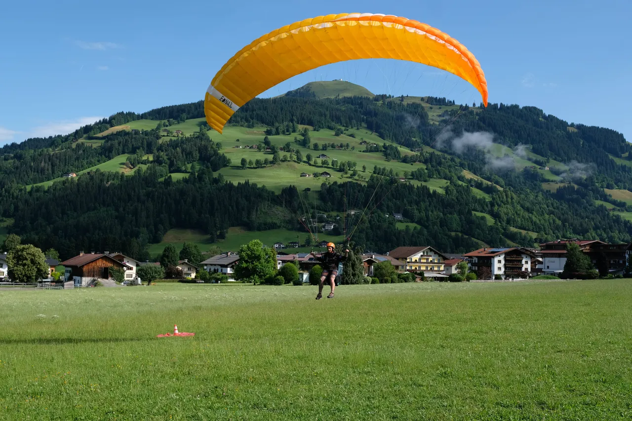 A paragliding student landing a yellow and orange canopy in a grassy field, with an Alpine village and forested hill in the background