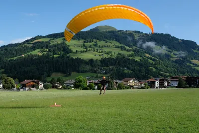 A paragliding student landing a yellow and orange canopy in a grassy field, with an Alpine village and forested hill in the background
