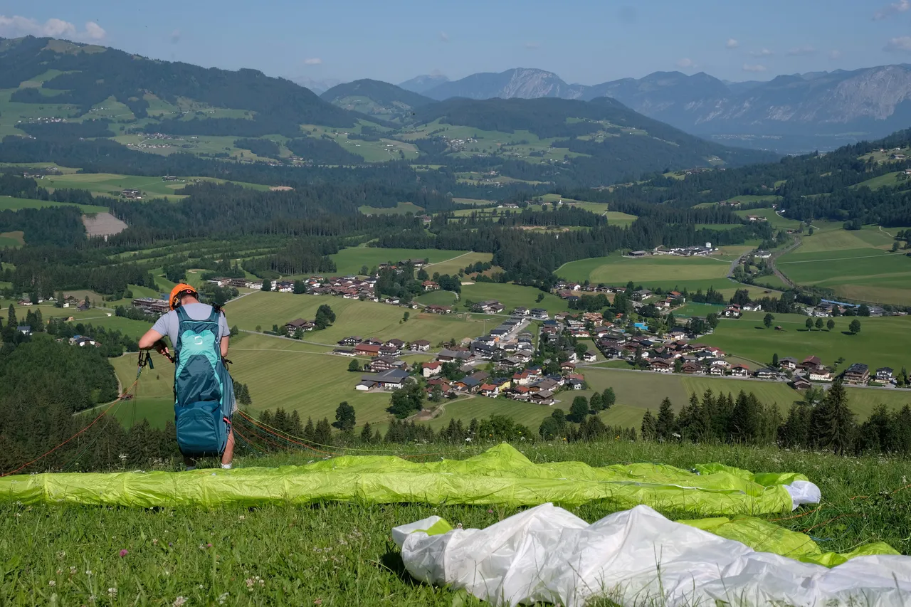 A paragliding student in helmet and harness standing at a launch site with the canopy laid out on the grass, overlooking a green alpine valley and village in Tyrol
