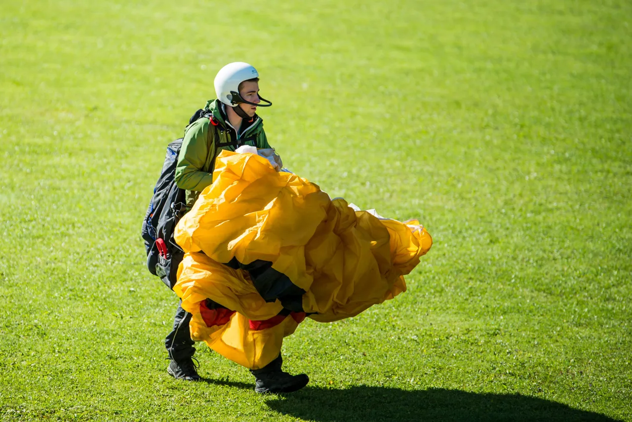 Paraglider pilot with helmet gathering up a yellow wing on a green meadow after landing
