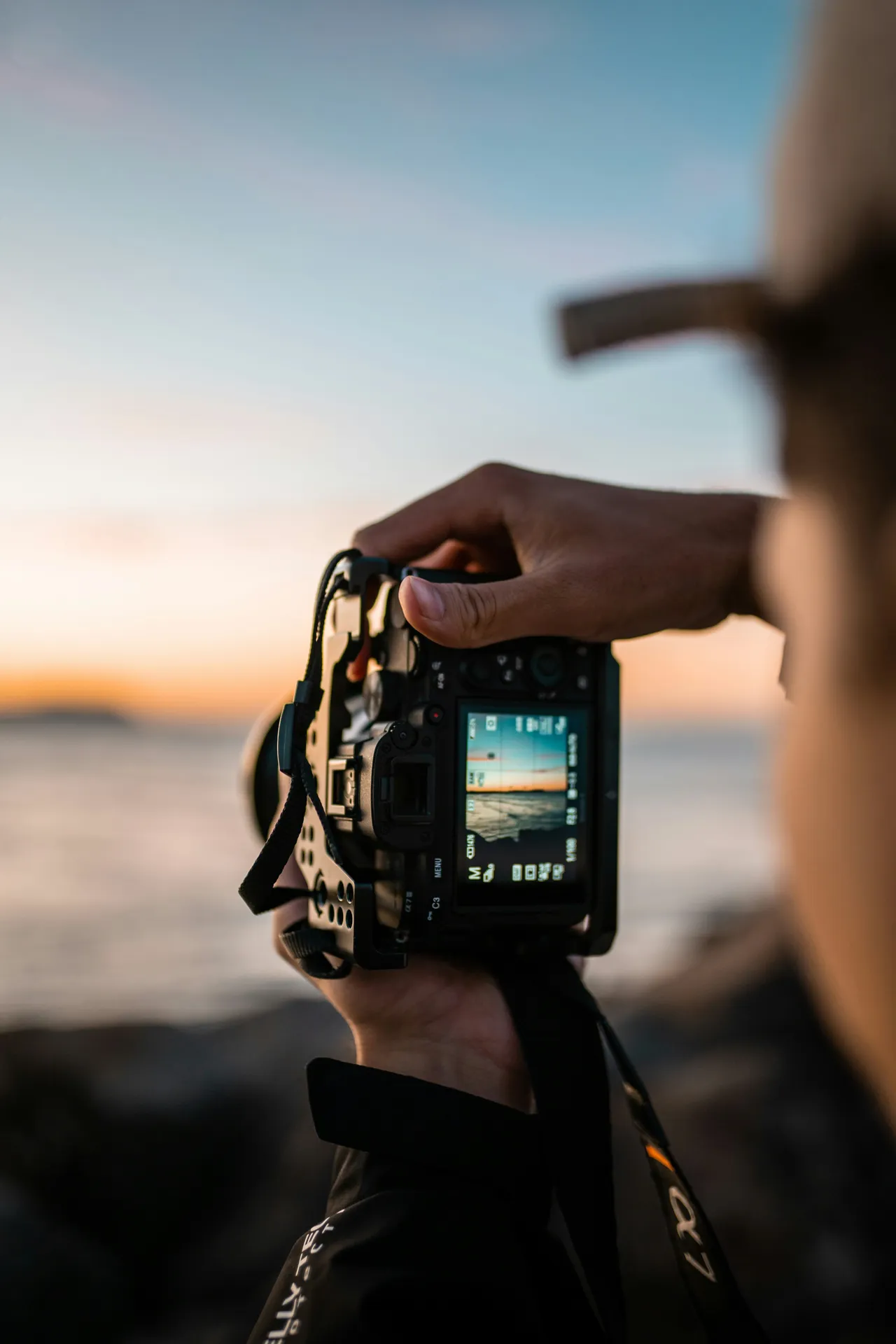 Person reviewing a sunset seascape photo on a camera screen by the ocean