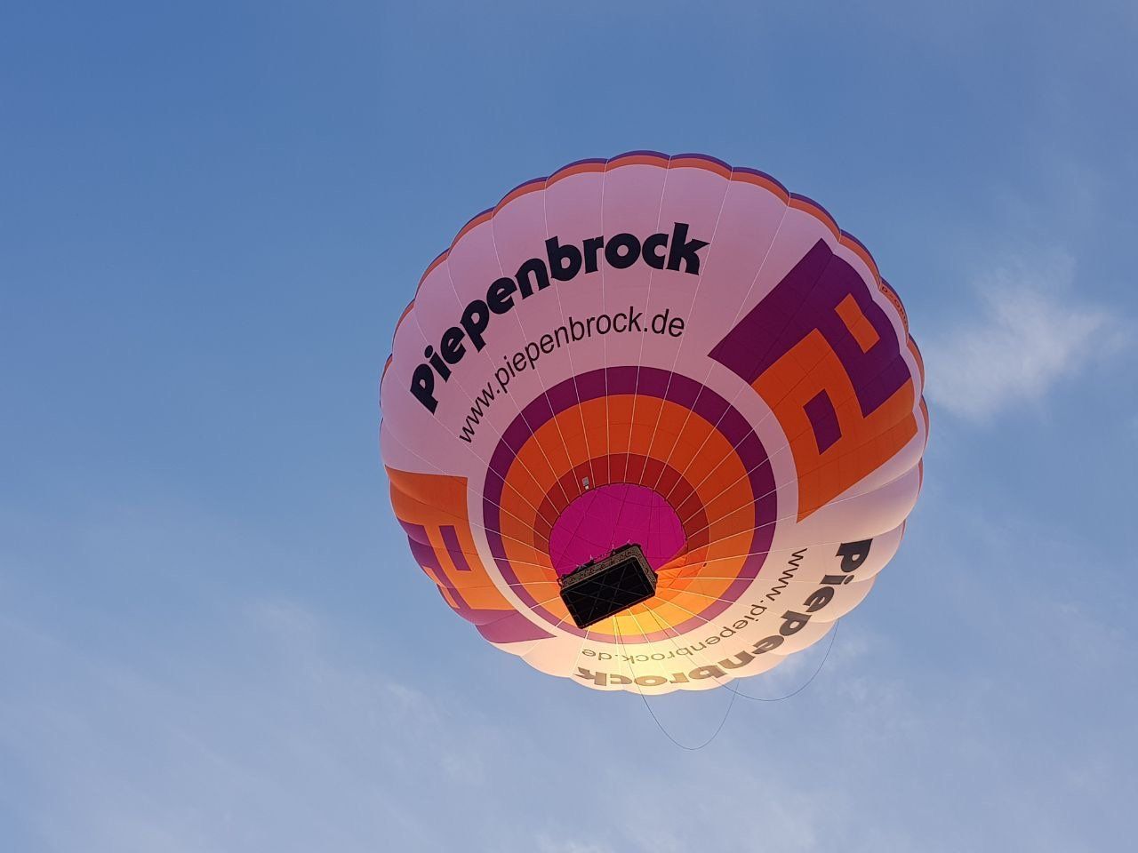 Looking up from directly below a pink, orange, and purple hot air balloon with Piepenbrock branding against a blue sky.