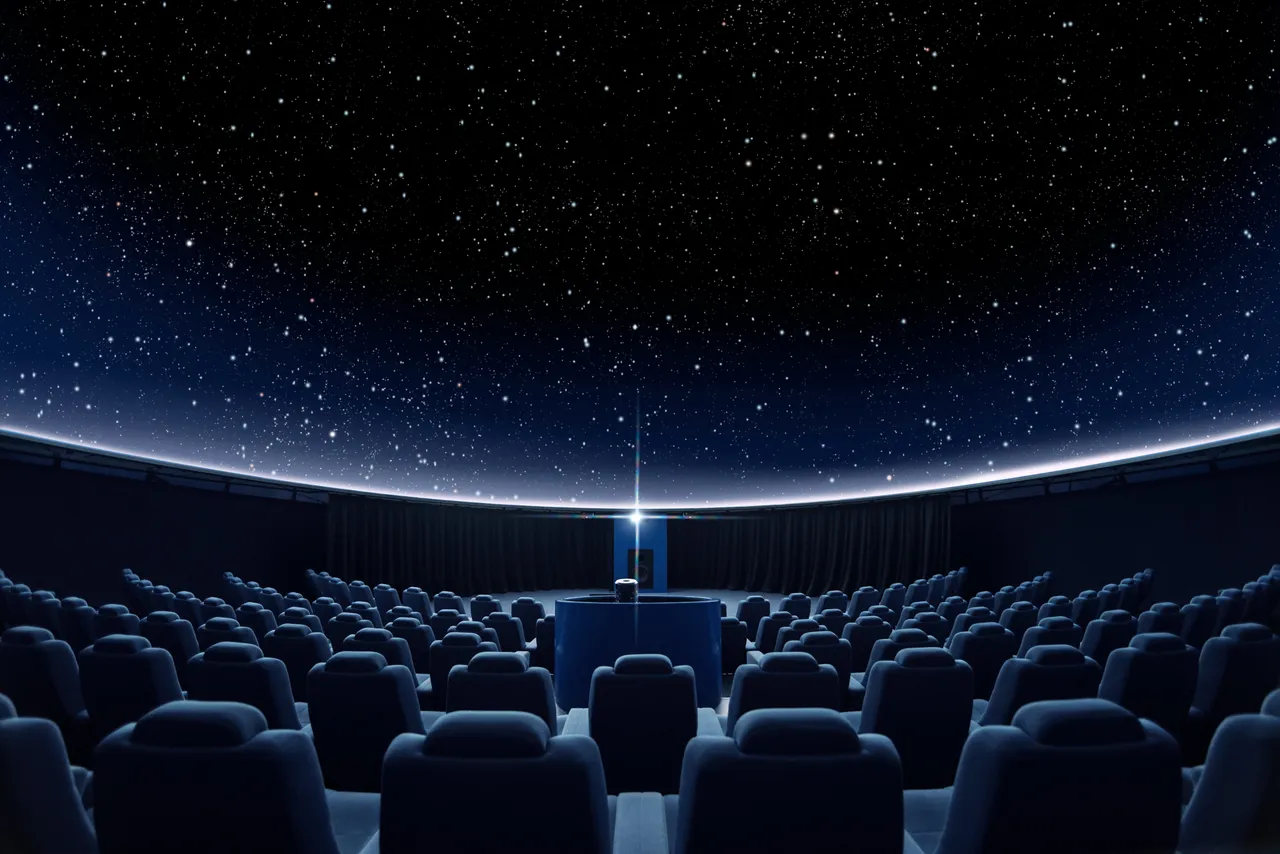 Symmetrical view of an empty planetarium theater showing blue rows of seating centered around a central projector casting a starry sky onto the domed ceiling.
