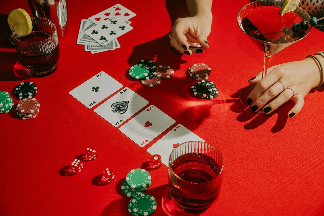 Four aces on a red poker table with chips, dice, and cocktails, hands with black nail polish