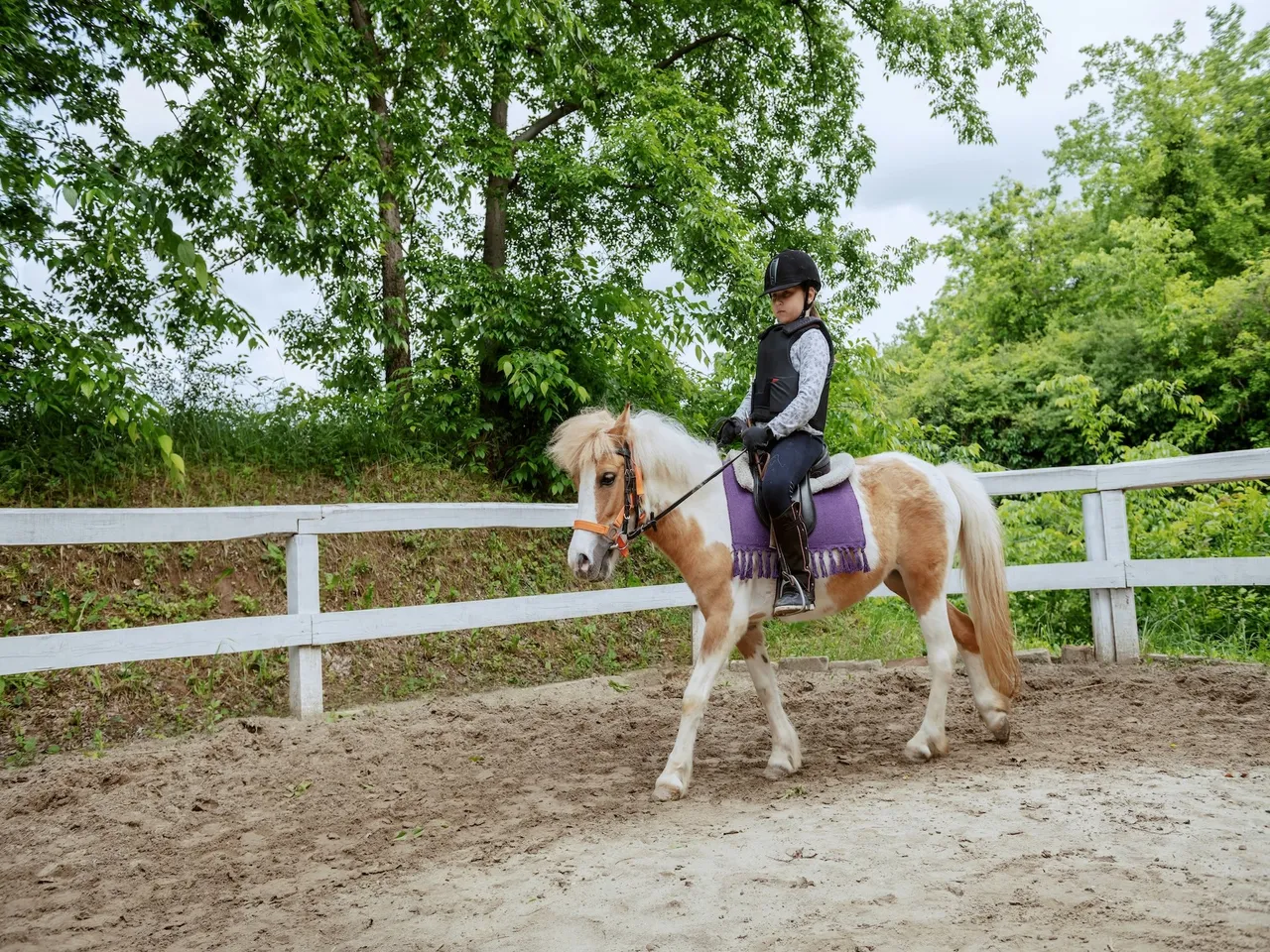 A young child wearing safety gear rides a small, light-brown and white pony along a wooden fence in a dirt arena.