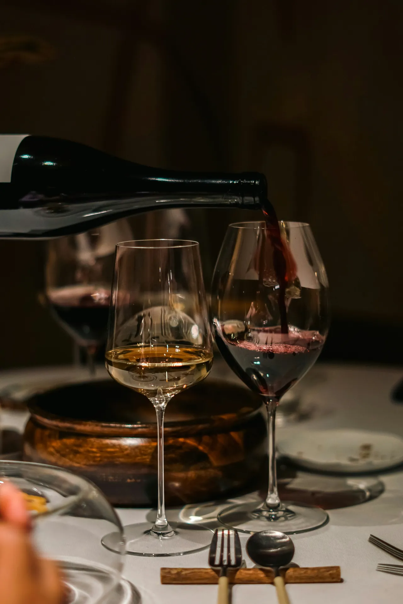 A close-up of red wine being poured from a dark green bottle into a wine glass on a set restaurant table next to a glass of white wine.
