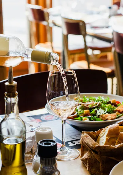 Rose wine being poured from a clear bottle into a wine glass on a restaurant table featuring a fresh salad, bread basket, and olive oil bottle.