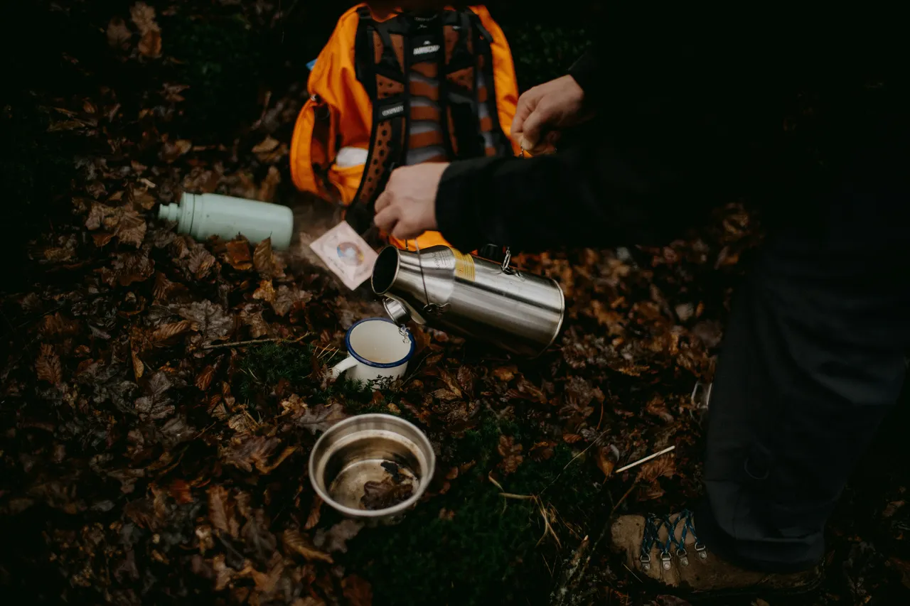 A person pouring hot water from a metal thermos into a mug on a forest floor covered in autumn leaves.