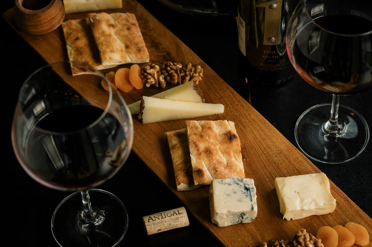 Two glasses of red wine sit alongside a long wooden charcuterie board featuring assorted cheeses, crackers, walnuts, and dried apricots.