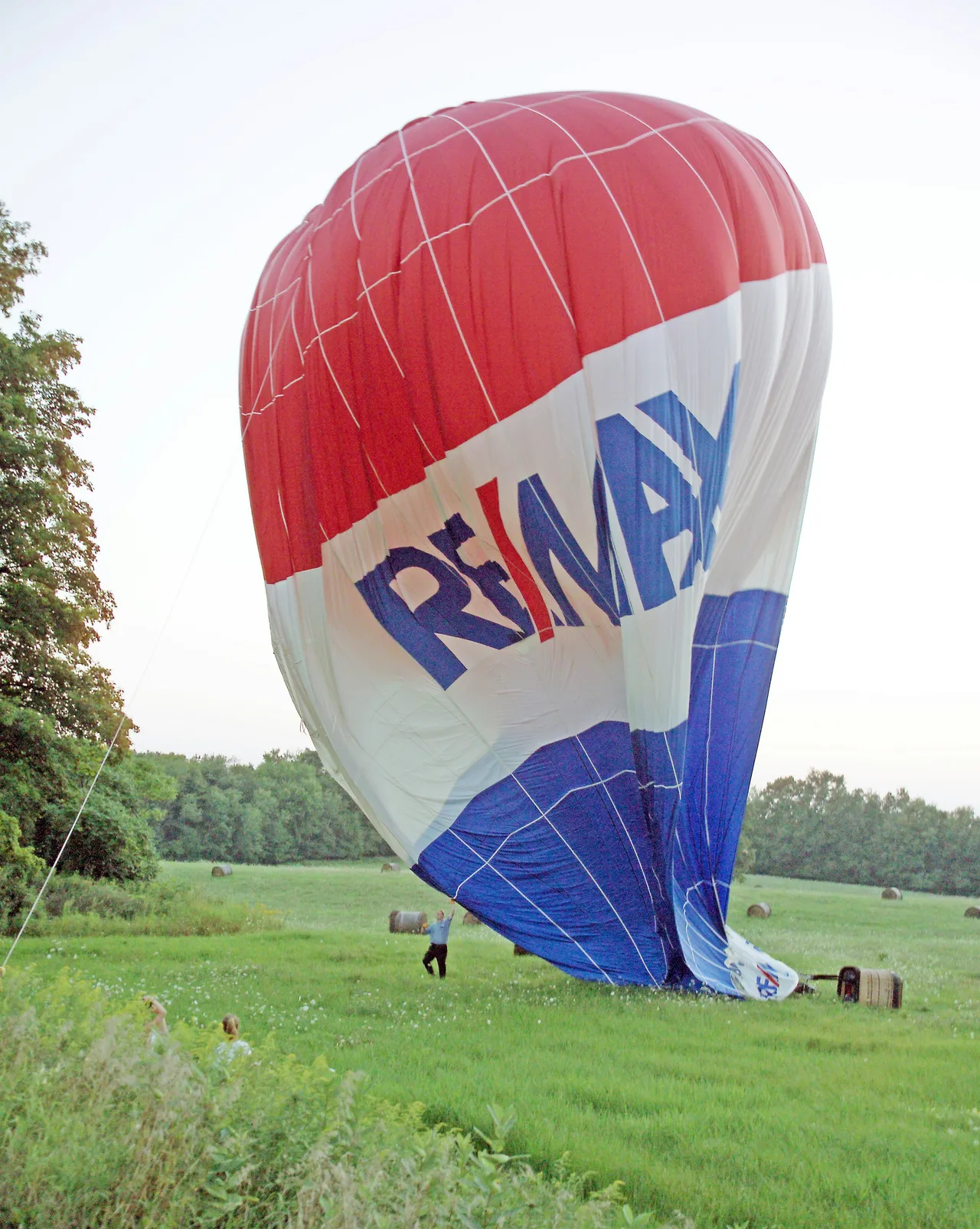 A red, white, and blue RE/MAX hot air balloon deflating on a green grassy field surrounded by trees.