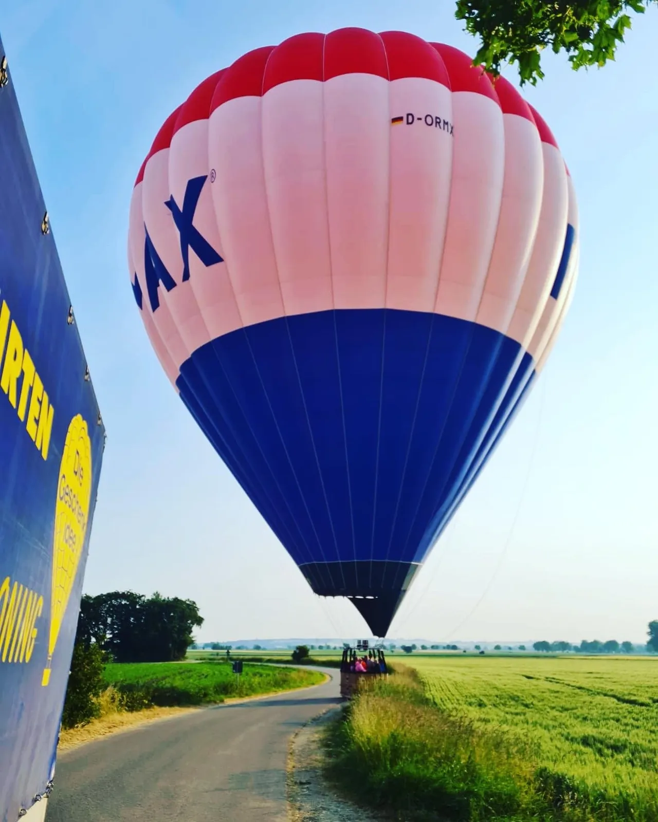 A large red, white, and blue RE/MAX hot air balloon preparing to land near a narrow rural road and green fields.