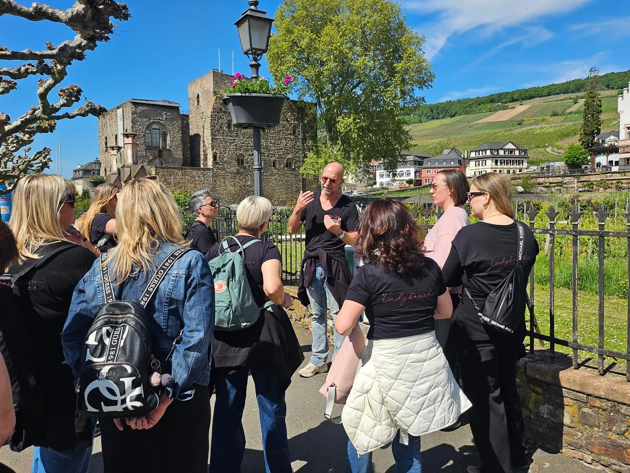 Tour guide talks to a group of women in matching black T-shirts in front of the medieval Brömserburg castle, with vineyards on the hills of Rüdesheim behind them.