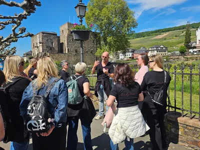 Tour guide talks to a group of women in matching black T-shirts in front of the medieval Brömserburg castle, with vineyards on the hills of Rüdesheim behind them.