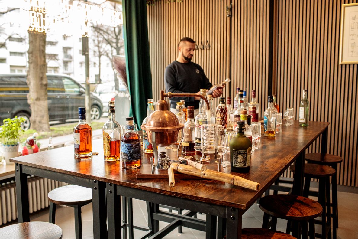 Host setting up a table featuring a small copper still, bamboo cane, and an array of rum bottles for a rum tasting.