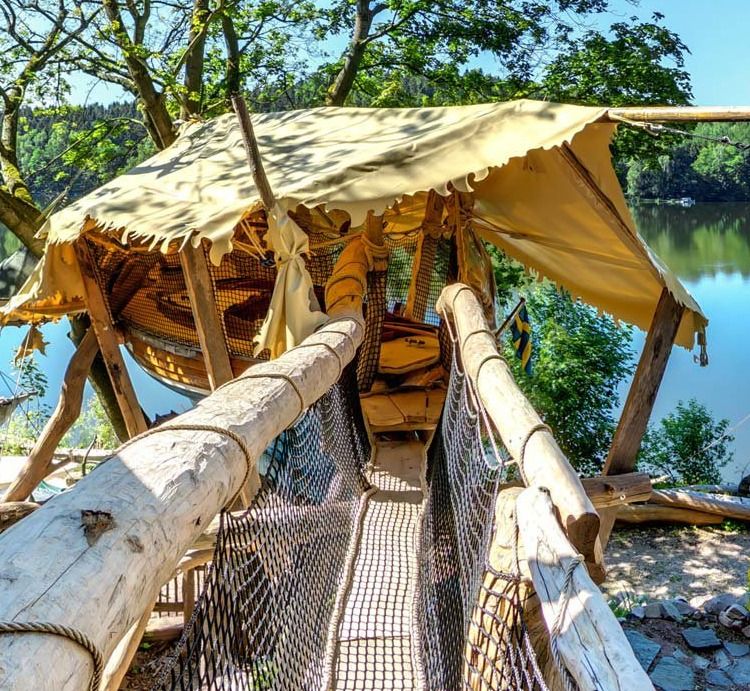 Rope bridge leading to a sailboat treehouse with canvas awning above the lake shore