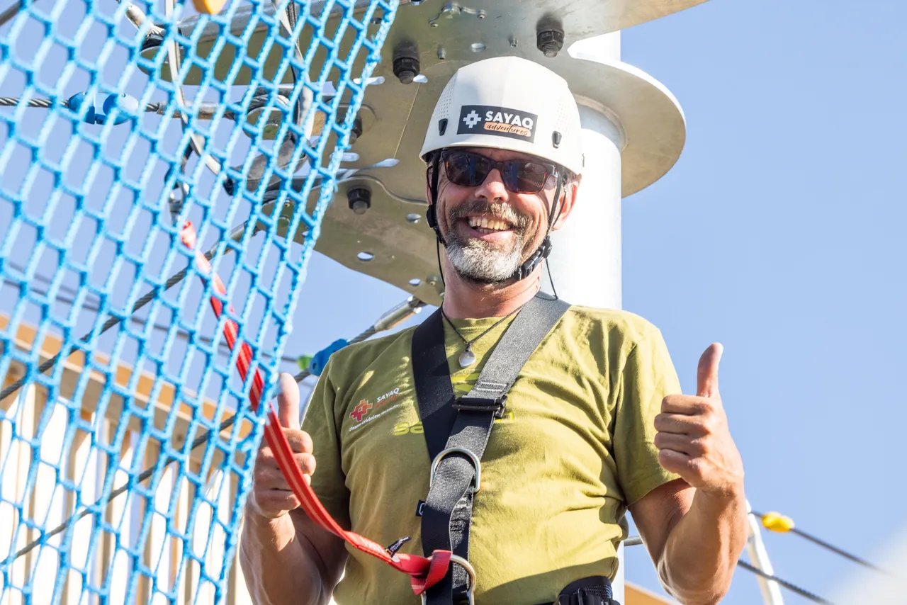 A smiling Sayaq Adventures climbing instructor wearing a white helmet, sunglasses, and a safety harness, giving a double thumbs-up at the Kristallturm ropes course.
