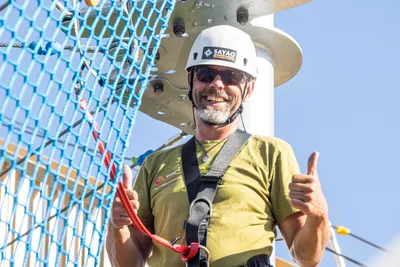 A smiling Sayaq Adventures climbing instructor wearing a white helmet, sunglasses, and a safety harness, giving a double thumbs-up at the Kristallturm ropes course.