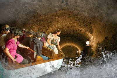 Visitors in VR headsets riding a boat through a stone tunnel alongside an actress in period Victorian dress.